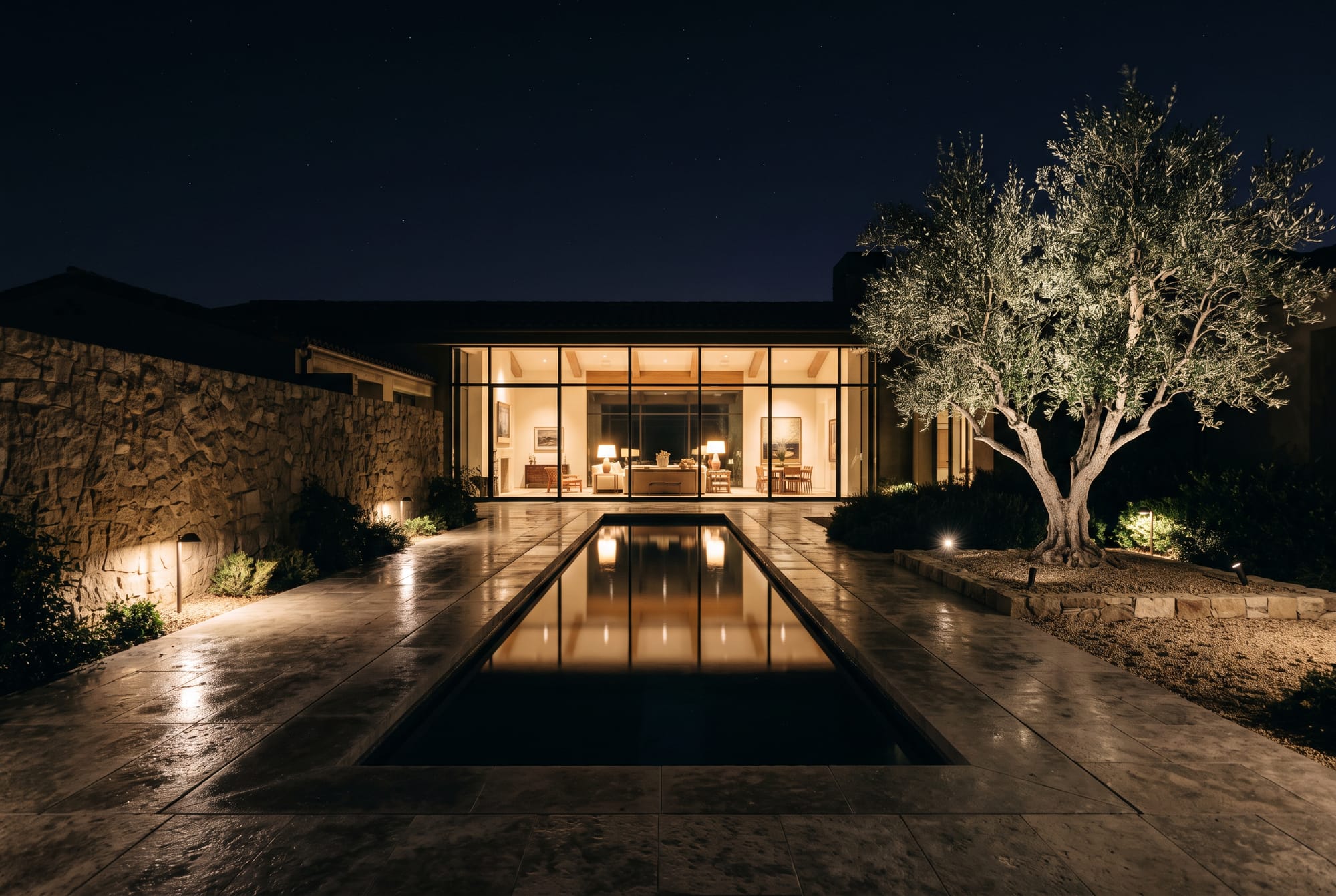 A courtyard at night, a single olive tree softly uplit beside a reflecting pool, the residence glowing beyond